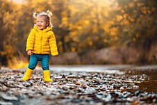 A Smiling Child Girl In Yellow Rubber Boots Is Playing On The River Bank. Copy Space. Concept Of Happy Childhood Stock Photo