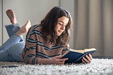 A Young Woman Is Reading A Book While Sitting On The Floor In Her Room. Stock Photos