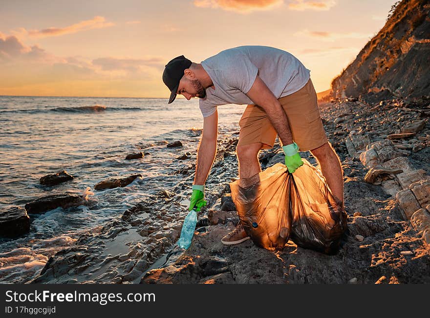 Cleaning beach from the pollution. Volunteer picking up a plastic bottle. In the background, the sea and the sunset