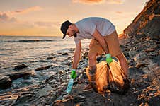 Cleaning Beach From The Pollution. Volunteer Picking Up A Plastic Bottle. In The Background, The Sea And The Sunset Stock Photo
