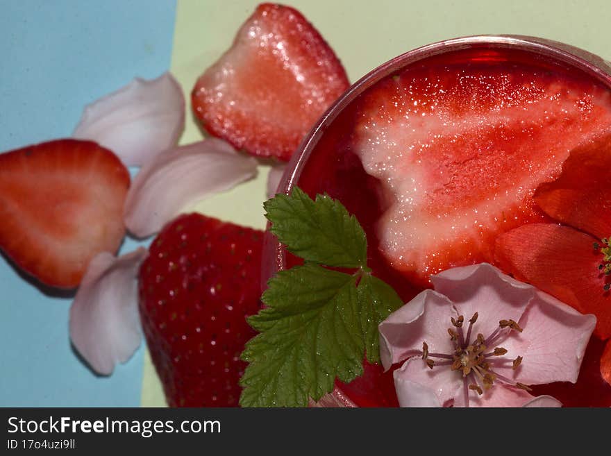 close-up and focus on a glass of strawberry juice with deokaricoja strawberry and flower head red and white under a glass of strawberry and white petal on a pastel green-blue backgroud, idyllic summer