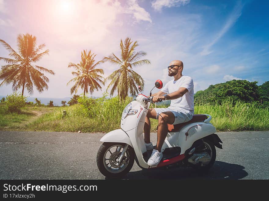 Stylish young man rides a motorbike on the road near the sea and palm trees.