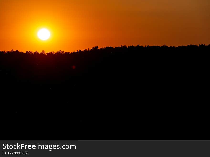 Beautiful sunset over the forest on a summer day. The dark silhouette of the forest and the bright blinding orange sun lightly touches the treetops on the horizon