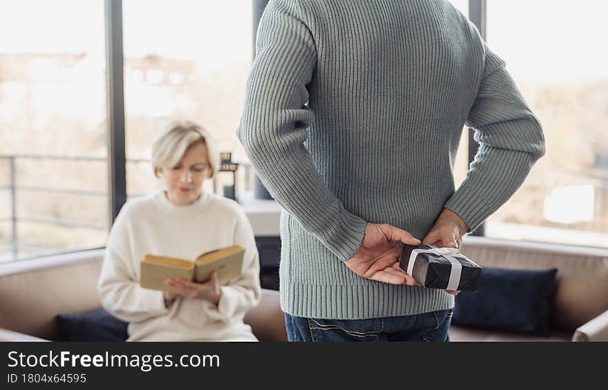 Cropped rear view of men holding gift box behind his back, while his wife sitting on couch and reading paper book. Surprise, birthday or anniversary concept. Cropped rear view of men holding gift box behind his back, while his wife sitting on couch and reading paper book. Surprise, birthday or anniversary concept