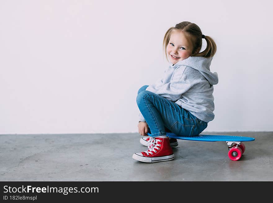 Smiling little girl in a summer dress fashion, sitting on a skateboard, and props up a head a hand, gazing at the camera with satisfaction and happiness. Positive emotions of children. Smiling little girl in a summer dress fashion, sitting on a skateboard, and props up a head a hand, gazing at the camera with satisfaction and happiness. Positive emotions of children