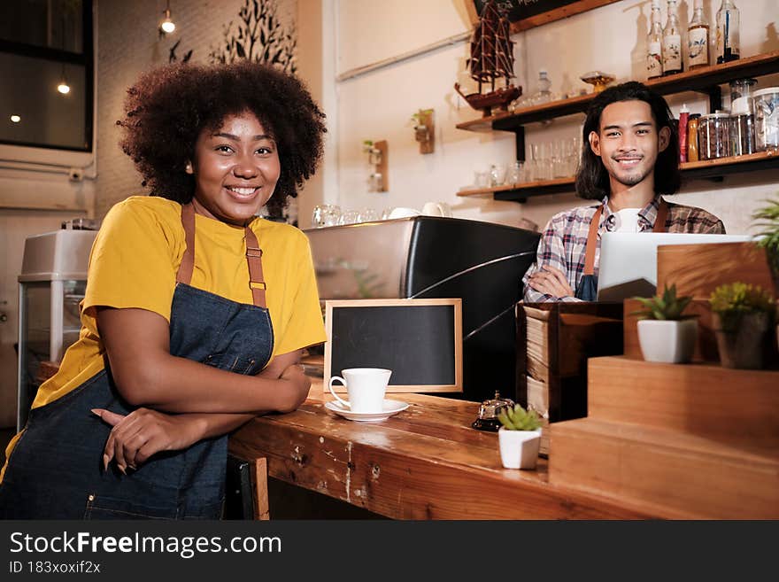Two cafe business startup partners and friends, African American female, and Thai male baristas look at camera, arms crossed, and cheerful smiles at coffee shop counter bar, happy service staff job. Two cafe business startup partners and friends, African American female, and Thai male baristas look at camera, arms crossed, and cheerful smiles at coffee shop counter bar, happy service staff job.