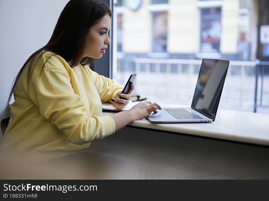 Young 20s woman digital nomad checking news from web pages and connecting smartphone to laptop. Young 20s woman digital nomad checking news from web pages and connecting smartphone to laptop