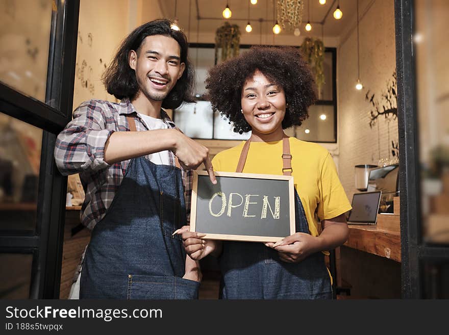 Two young startup barista partners with aprons stand at casual cafe door, letters on board and show open sign, happy and cheerful smiles with coffee shop service jobs, and new business entrepreneurs. Two young startup barista partners with aprons stand at casual cafe door, letters on board and show open sign, happy and cheerful smiles with coffee shop service jobs, and new business entrepreneurs.