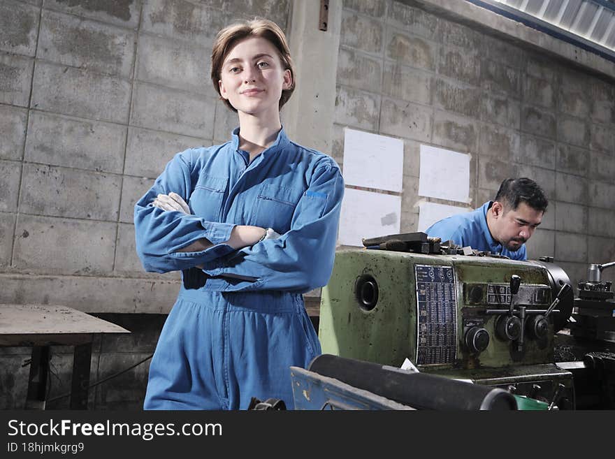 Portrait of Young White female industry worker stands, arms crossed, looks at camera and smiles, male partner works with machine behind her in manufacturing factory, professional mechanical engineers. Portrait of Young White female industry worker stands, arms crossed, looks at camera and smiles, male partner works with machine behind her in manufacturing factory, professional mechanical engineers.