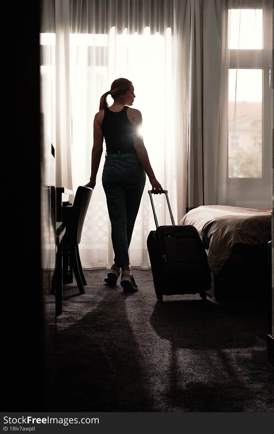 Woman traveler with luggage suitcase after checking Into a Hotel Room looking in the window. Silhouette of visitor in travel accommodation ready to have rest after arrival. Woman traveler with luggage suitcase after checking Into a Hotel Room looking in the window. Silhouette of visitor in travel accommodation ready to have rest after arrival