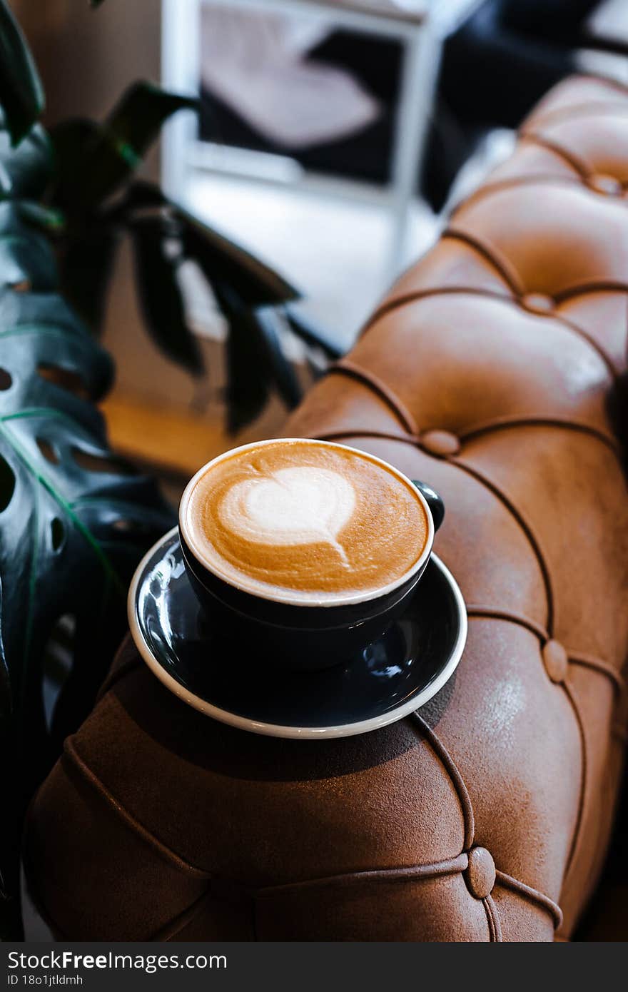 Black cup of cappuccino with latte art on wooden background. Beautiful foam, green monstera plant at cafe