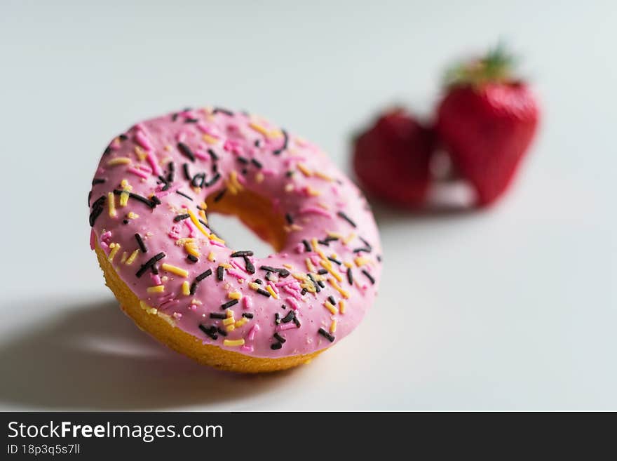 A photo of a donut with pink icing, a small part of which is in focus and a strawberry in the background, very out of focus. All objects on a light background