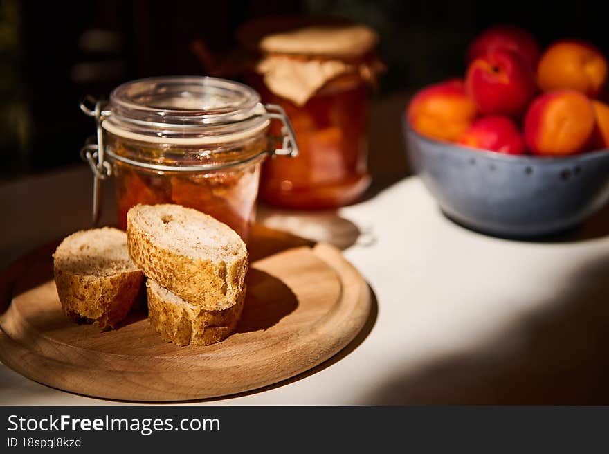 Still life, illuminated by a daylight. Close-up slices of whole grain bread on wooden board, jar with homemade jam and ripe juicy apricots in blue ceramic bowl on white table in rustic summer kitchen. Still life, illuminated by a daylight. Close-up slices of whole grain bread on wooden board, jar with homemade jam and ripe juicy apricots in blue ceramic bowl on white table in rustic summer kitchen