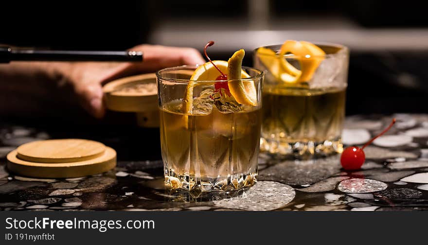 A narrow view of hands preparing Old Fashioned cocktails with smoker. Selective focus on front drink.