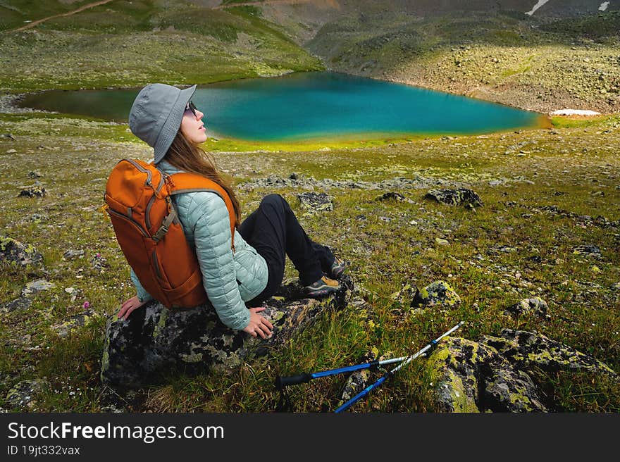 nice woman in a panama with a backpack sits on a stone, having a rest after trekking in the mountains, near the lake.