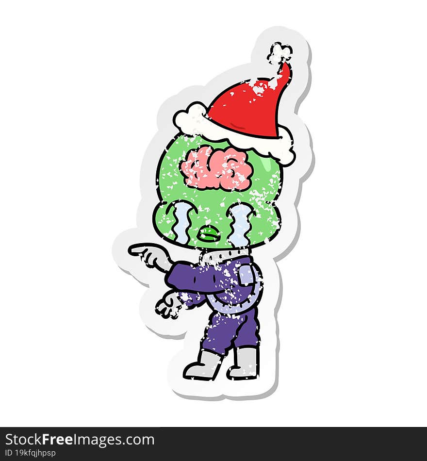 distressed sticker cartoon of a big brain alien crying and pointing wearing santa hat