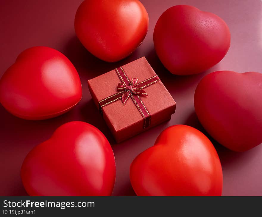 A small red present gift box with ribbon surrounded with many heart balls on red background. An essential gift with on special days with love, birthdays, New Years, Valentine`s Day and anniversaries.