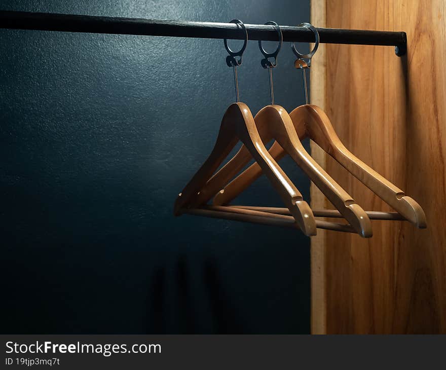 Three empty wooden clothes hangers without shirts or dress hanging on a black cloth rack in the wood wardrobe on dark blue background preparing for the guest who staying in the room in the hotel. Three empty wooden clothes hangers without shirts or dress hanging on a black cloth rack in the wood wardrobe on dark blue background preparing for the guest who staying in the room in the hotel.