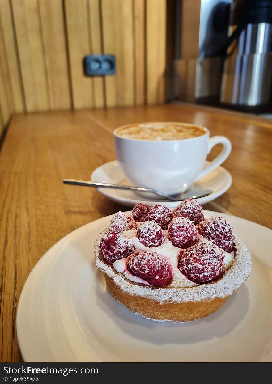 Raspberry tart on a white saucer in front of a cup of coffee