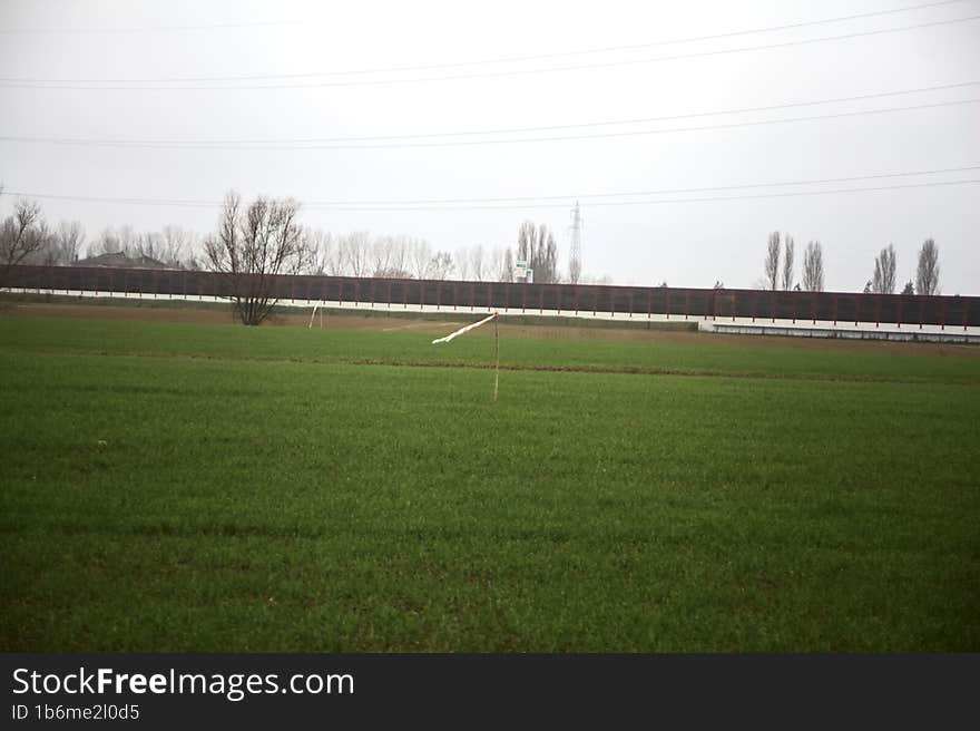 Cultivated field with trees in the distance framed by wooden pylons and over head cables on a cloudy day