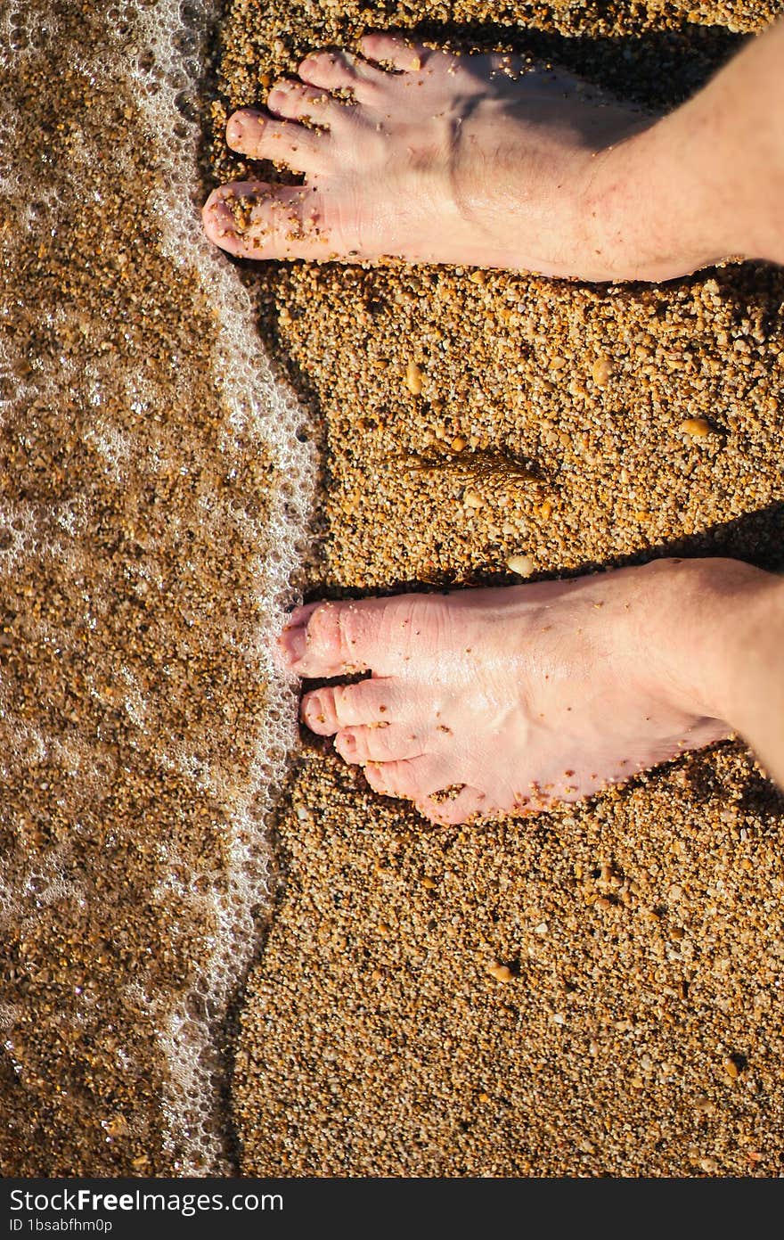 Men s feet are standing on a sandy beach. Small waves are foaming on the seashore. Crystal clear water.