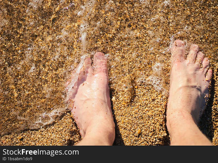 Men s feet are standing on a sandy beach. Small waves are foaming on the seashore. Crystal water.
