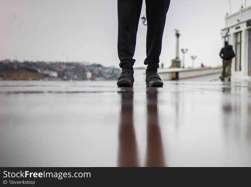 A man stands on wet asphalt in the rain. A park on the seashore. A muddy reflection on the ground.