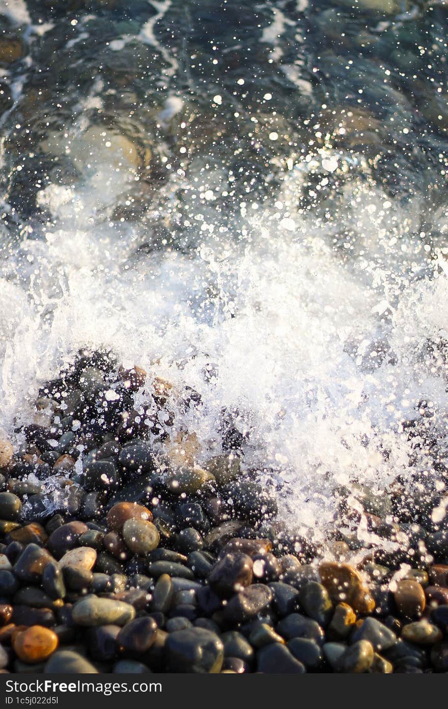 Splashing waves on the seashore. The beach is made of small colorful pebbles. Sea water splashes hitting the stones.