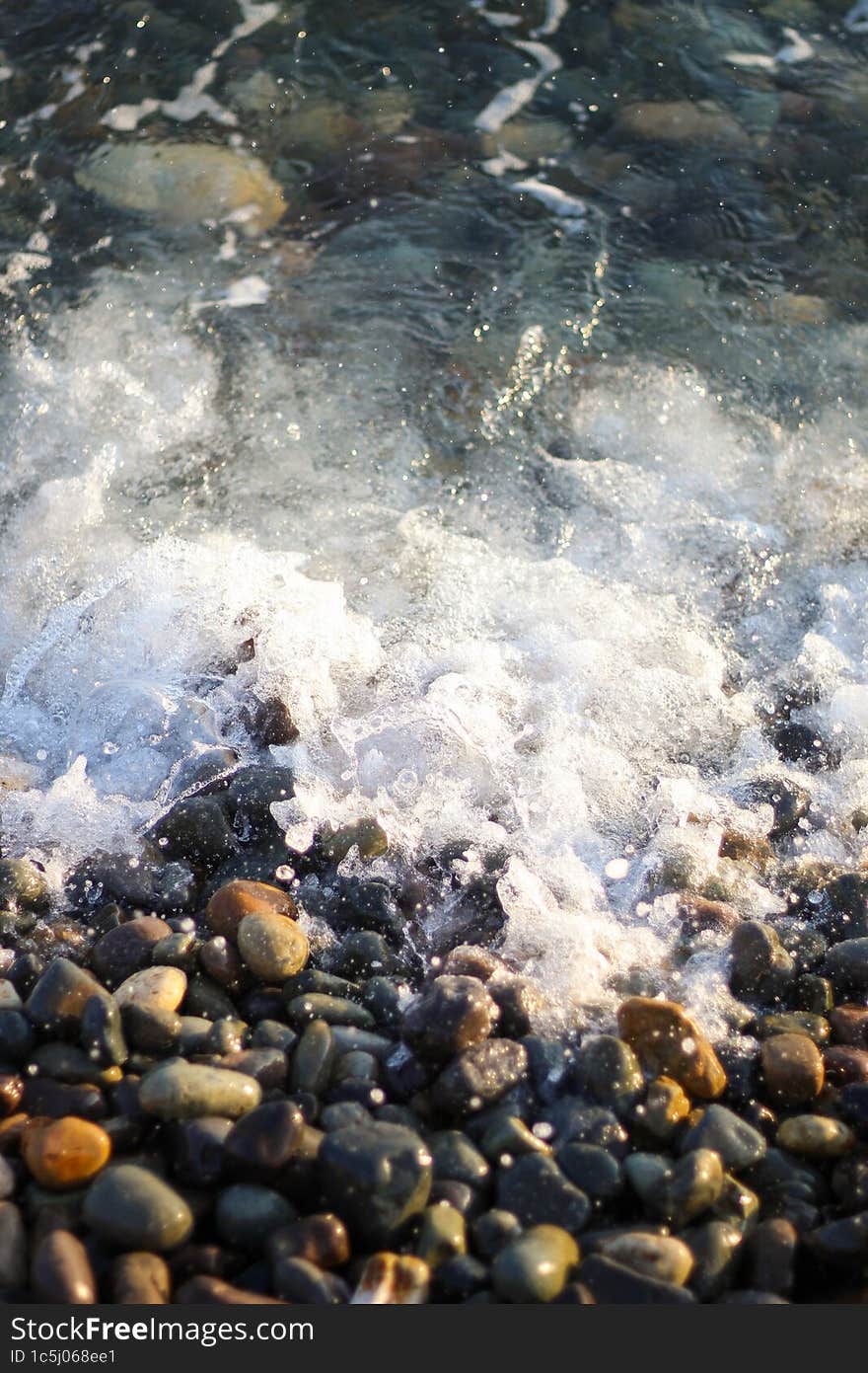 Splashing waves on the seashore. The beach is made of small colorful pebbles. Sea water splashes hitting the stone