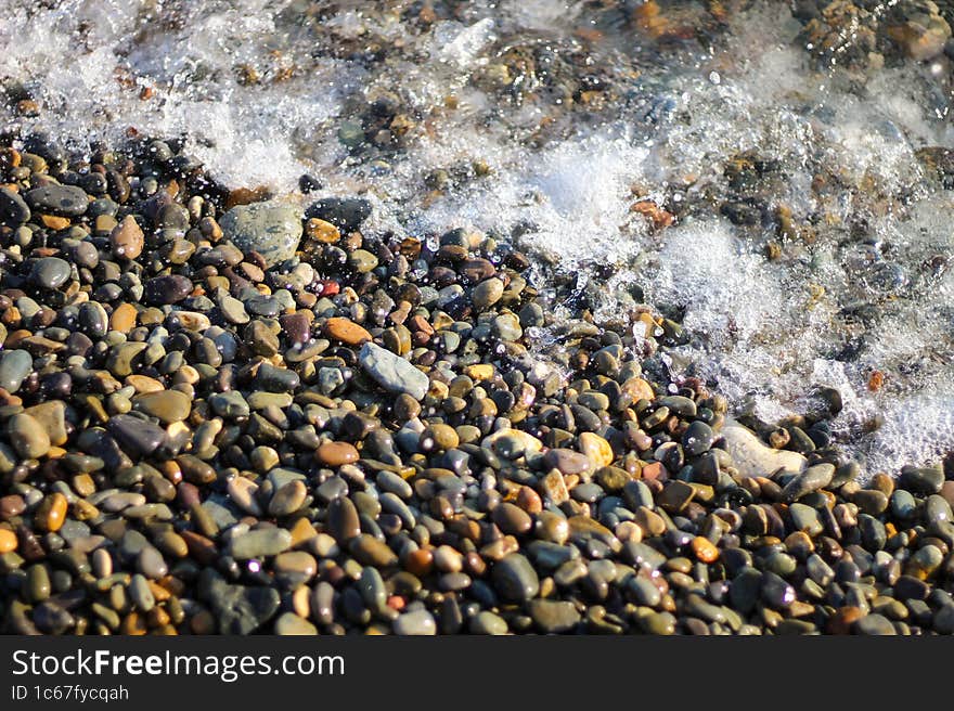 Splashing Waves On The Seashore. The Beach Is Made Of Small Colorful Pebbles. Sea Water Splashes Hitting The Stones.