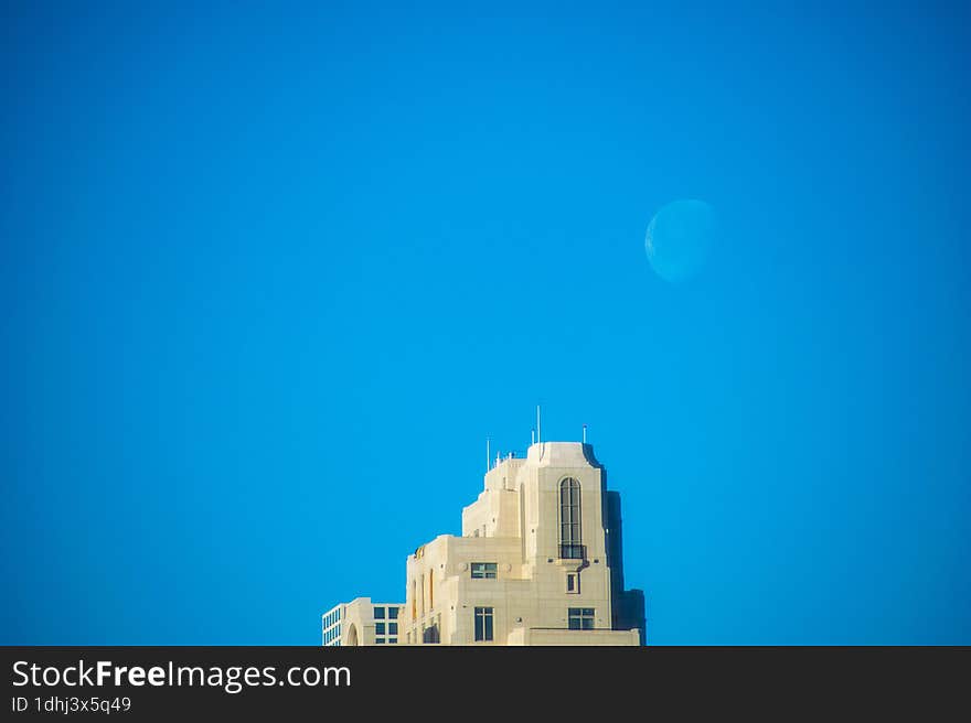 A bright white crescent moon rises over a blocky tan stone high rise skyscraper building with a true blue sky