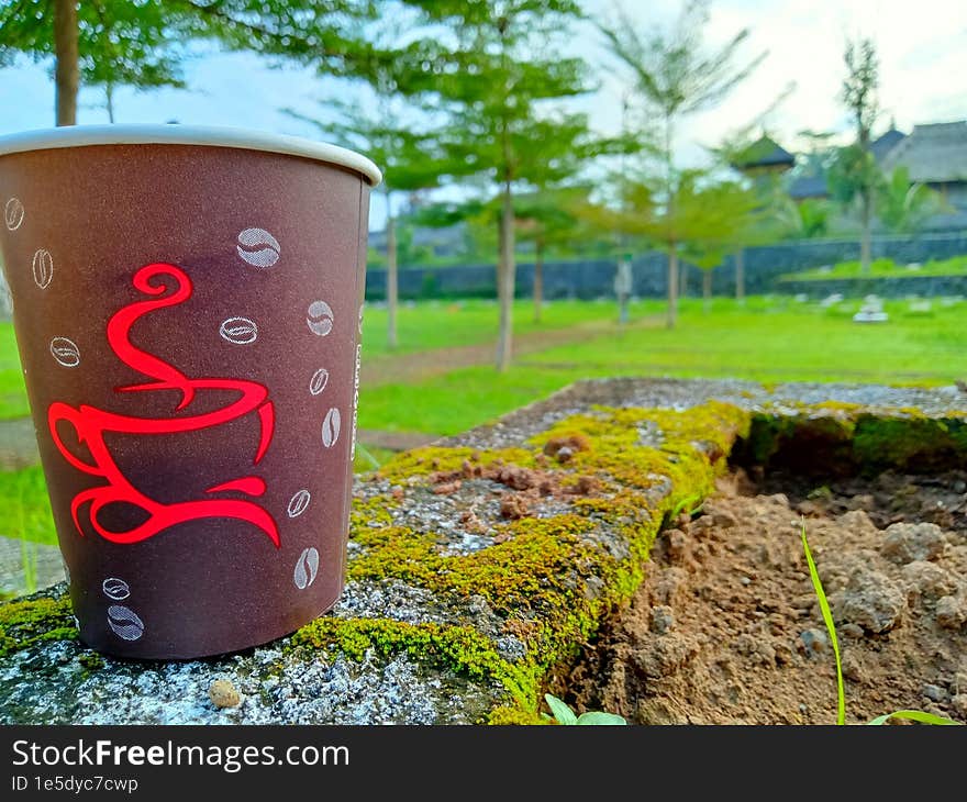 Close up photo of a paper cup filled with coffee against a city park background in the morning