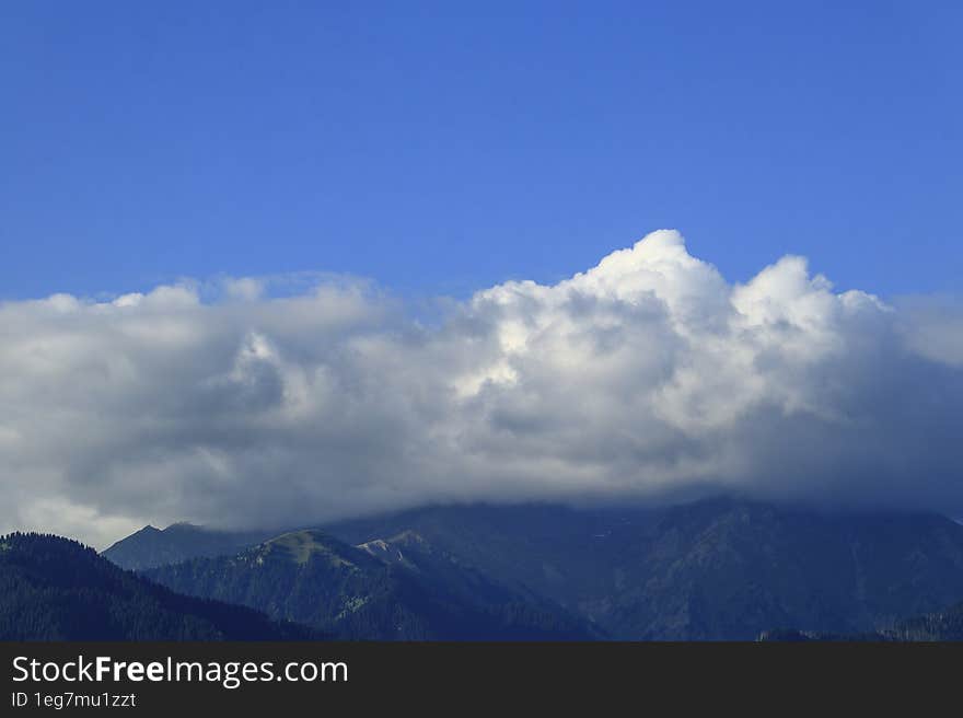 Large great white cumulus clouds in the blue sky over the mountains of the Trans-Ili Alatau, covering their peaks, are brightly li