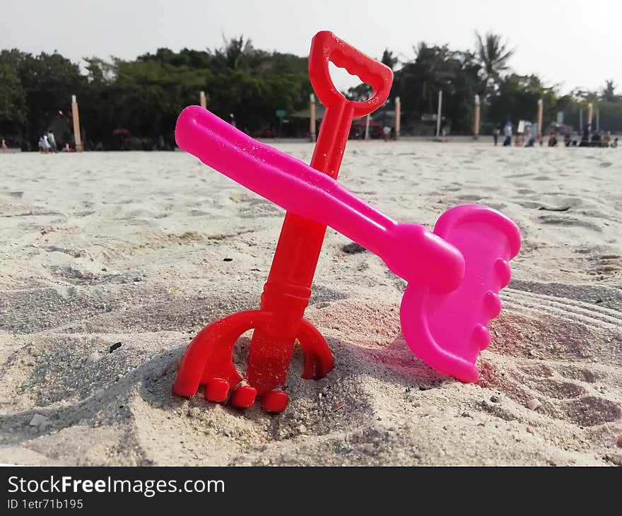 Red and pink sand rake toys in beach sand and people background taken in Jakarta Indonesia