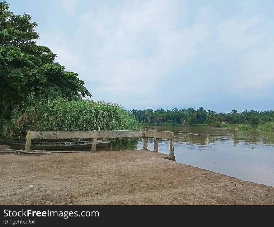 picture of a small harbor on the river bank with very murky water and from afar you can see an island with an oil palm plantation