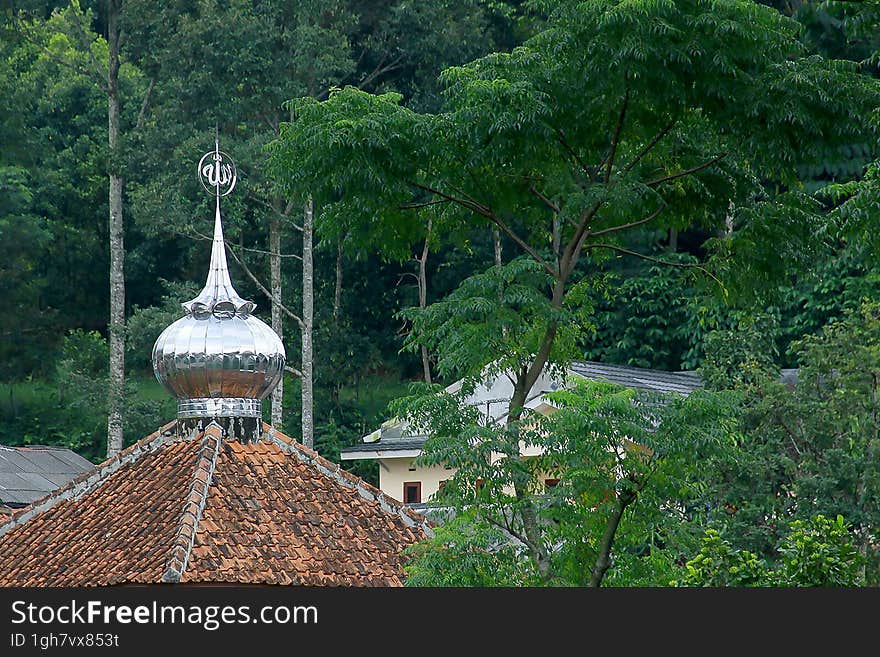 Image of the tile roof of a small mosque, with a small aluminum dome on top, in the middle of green forest trees.