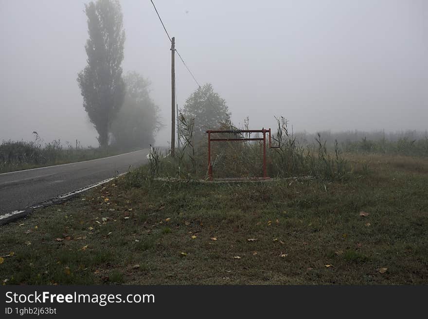 Narrow road bordered by a few trees and trenches with weirs on a foggy day in the italian countryside