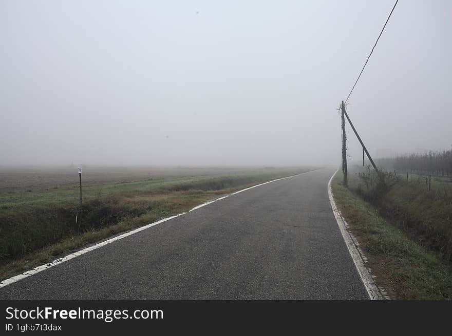 Road bordered by a trench and a power line next to fields on a foggy day in the italian countryside