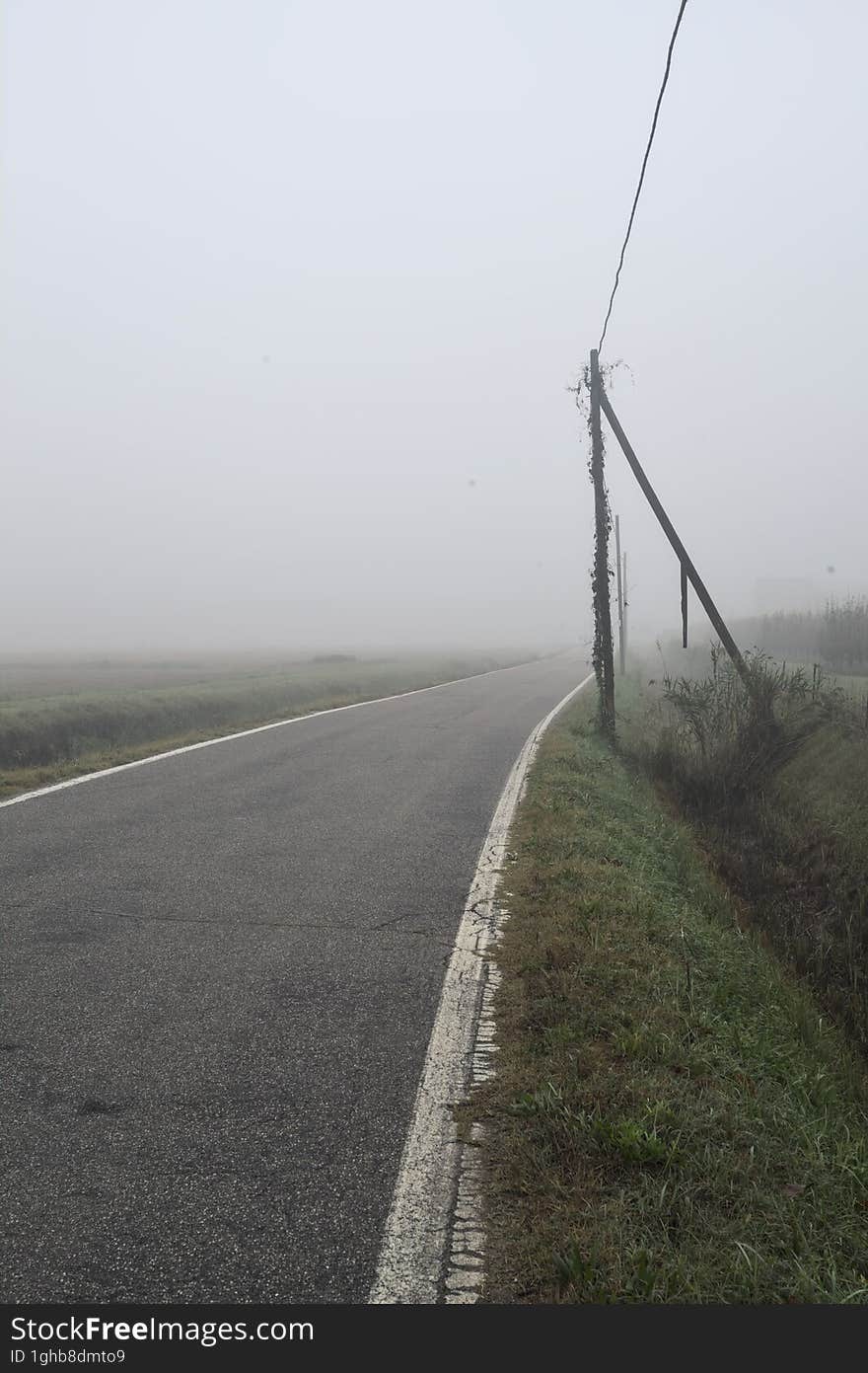 Road bordered by a trench and a power line next to fields on a foggy day in the italian countryside