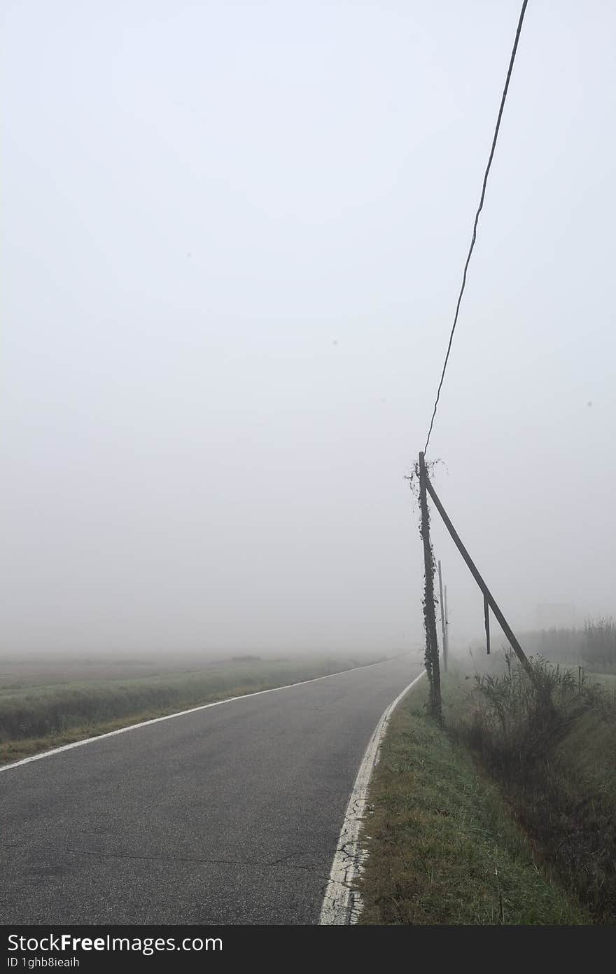 Road bordered by a trench and a power line next to fields on a foggy day in the italian countryside