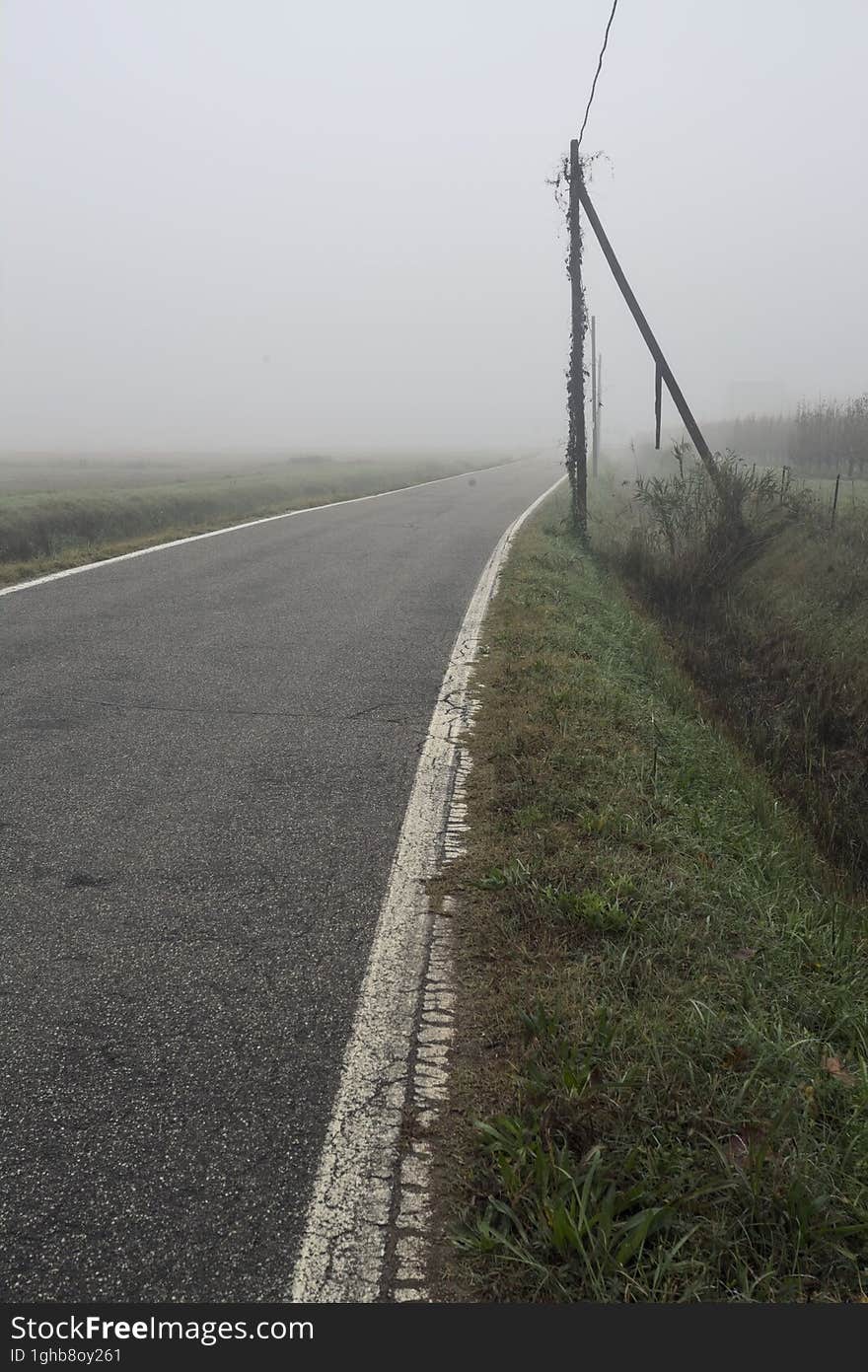 Road bordered by a trench and a power line next to fields on a foggy day in the italian countryside