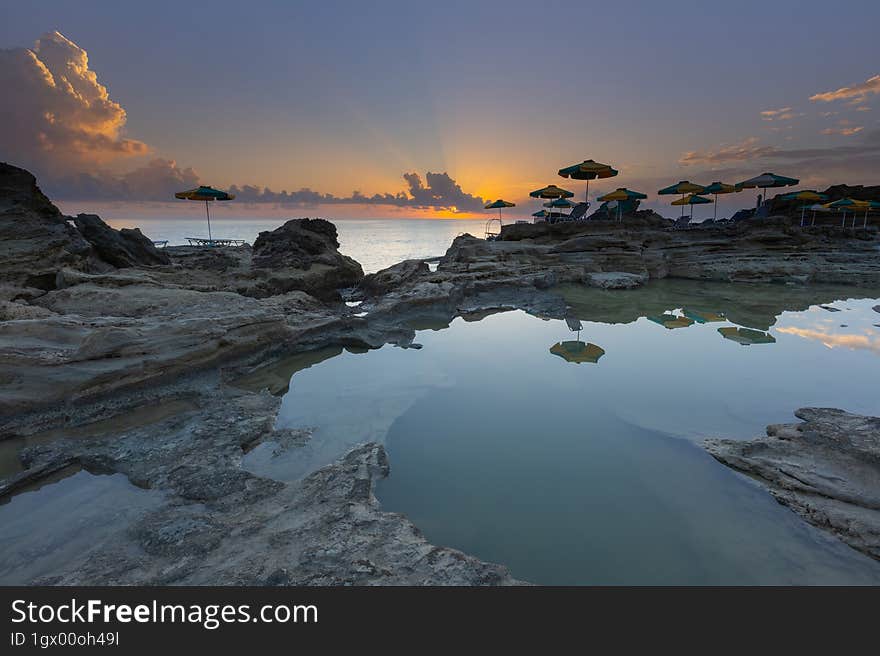 A romantic bay by the sea at sunrise with small lagoons in which the sky is reflected. Among the lagoons, there are umbrellas with sunbeds on the stones