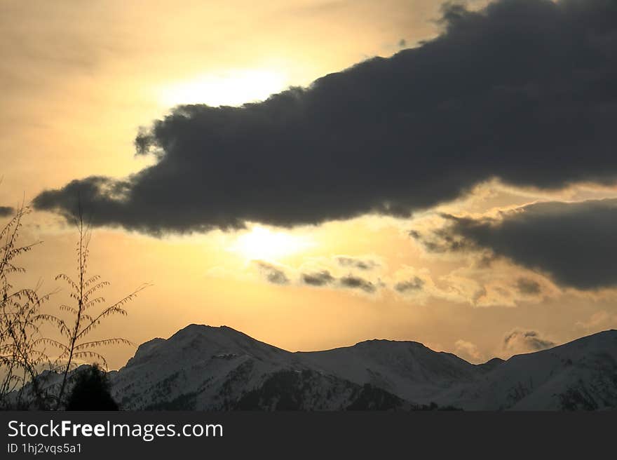 The sun behind a dark cloud over the snow-capped mountains of the Trans-Ili Alatau during sunrise behind thickets of autumn trees