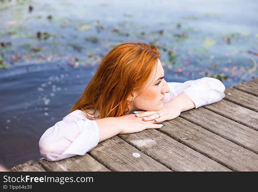 Red hair woman in swimsuit relaxing on wooden bridge near lake.