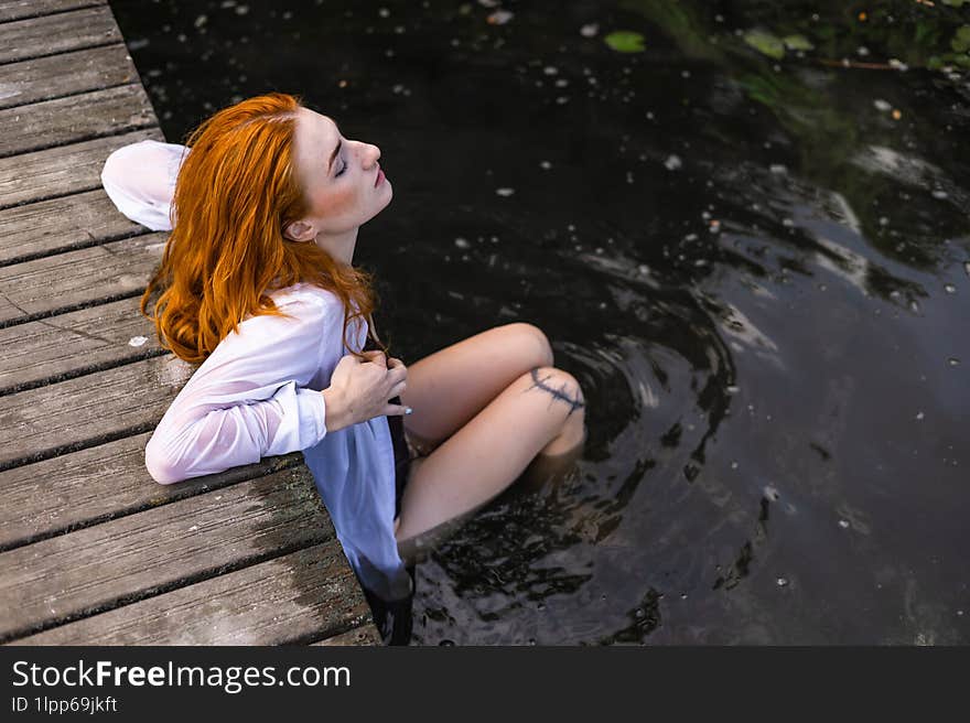 Red Hair Woman In Swimsuit Relaxing On Wooden Bridge Near Lake.