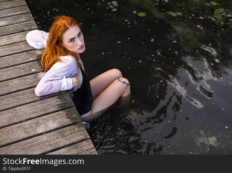 Red hair woman in swimsuit relaxing on wooden bridge near lake.