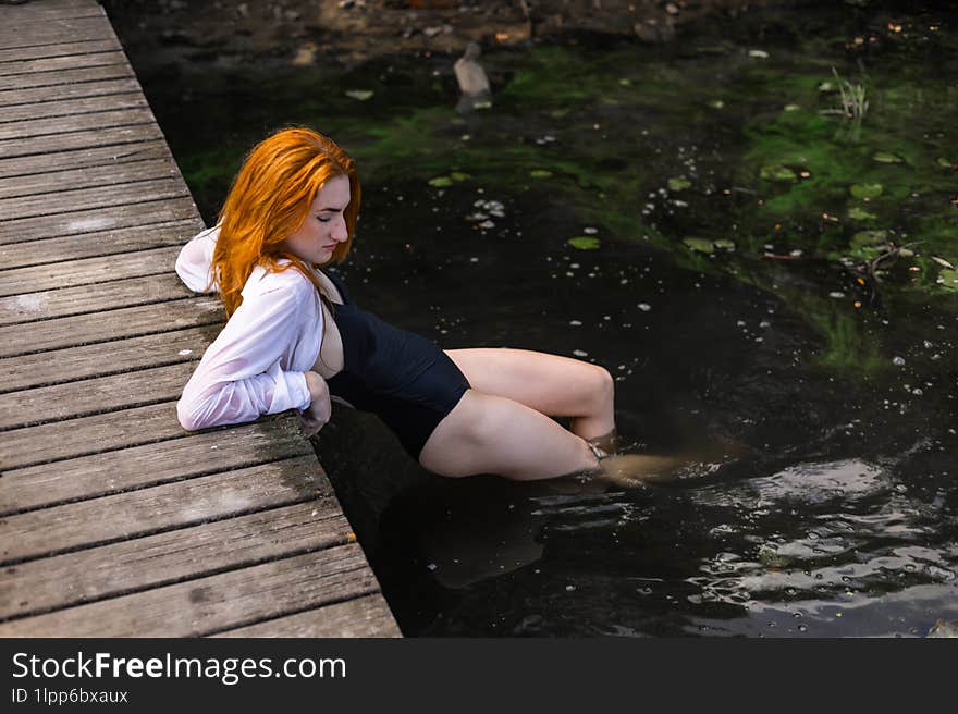 Red hair woman in swimsuit relaxing on wooden bridge near lake.