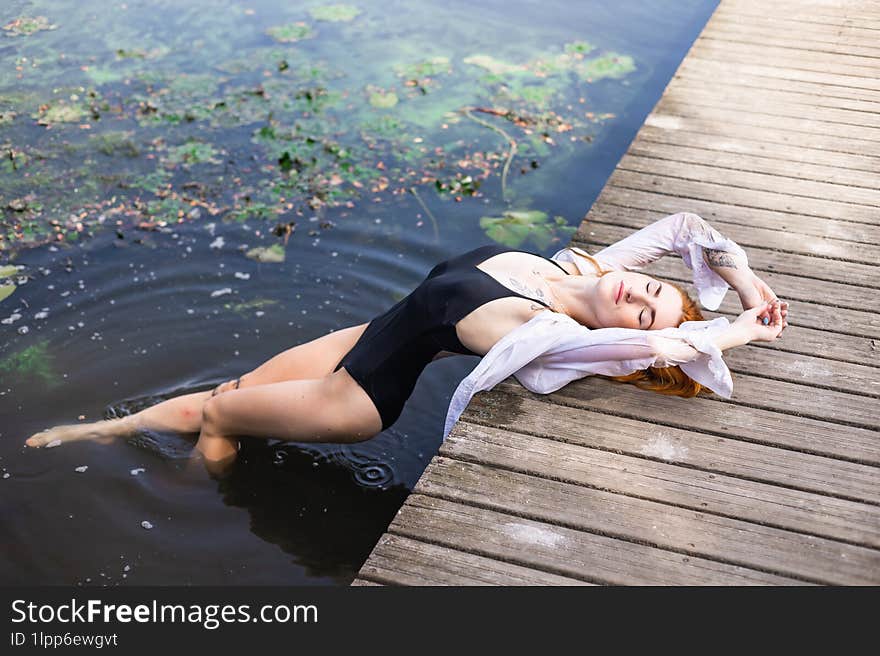 Red hair woman in swimsuit relaxing on wooden bridge near lake.