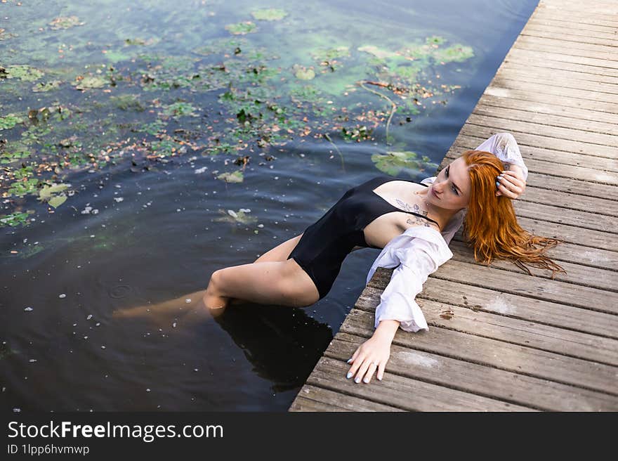 Red Hair Woman In Swimsuit Relaxing On Wooden Bridge Near Lake.