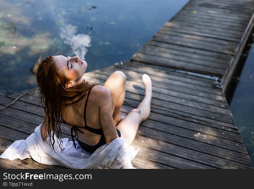 Red hair woman in swimsuit smoking on wooden bridge near lake.